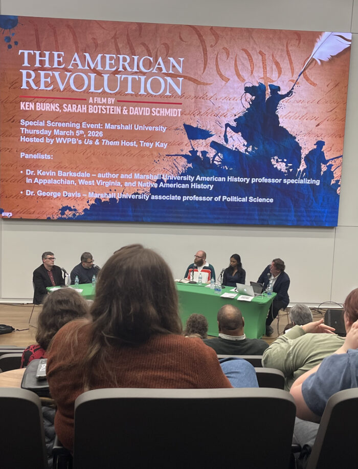 A photo at a distance showing all panelists for a panel discussion sitting at a table with a green tablecloth. Above them is a poster for Ken Burns' The American Revolution.