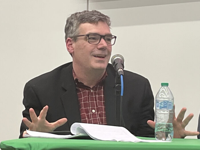A man with salt and pepper hair and glasses speaks on a panel. He wears a black jacket and red checkered shirt.