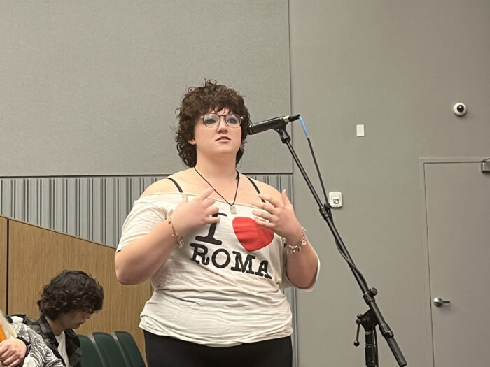 A young person with curly hair and wears glasses speaks at a standing microphone from the audience. She wears a shirt that says "I heart Roma."