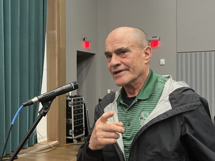 An older, balding man stands at a standing mic and speaks to panelists and the audience.