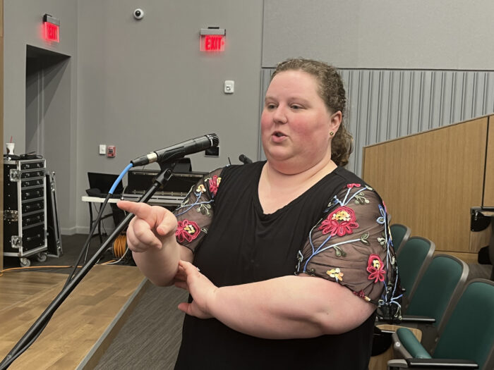 A woman with curly hair pulled back into a low ponytail. She wears a black dress shirt with flowers along the sleeve. She speaks at a standing mic.