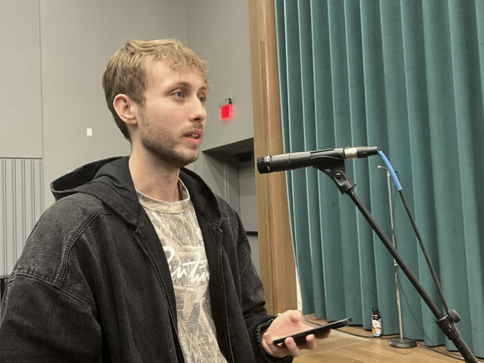 A younger man wearing a t-shirt and black jacket, stands at a standing microphone and speaks to a panel.