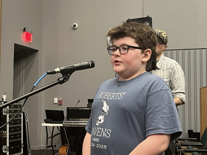 A young boy stands at a standing microphone and speaks to a panel of speakers. The boy has brown hair, wears glasses, and a blue t-shirt.