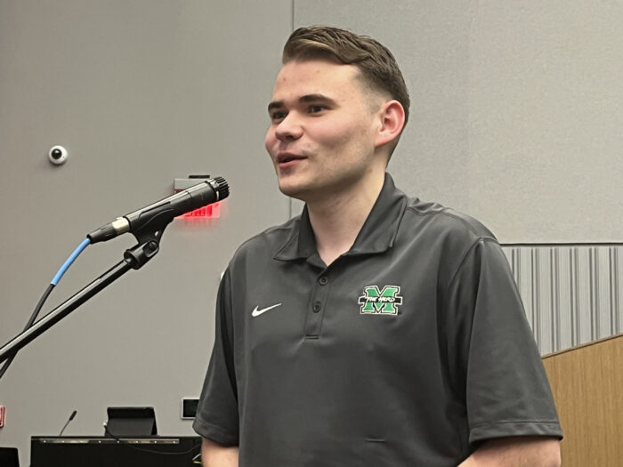 A male college student stands at a standing microphone and asks questions of a group of panelists.