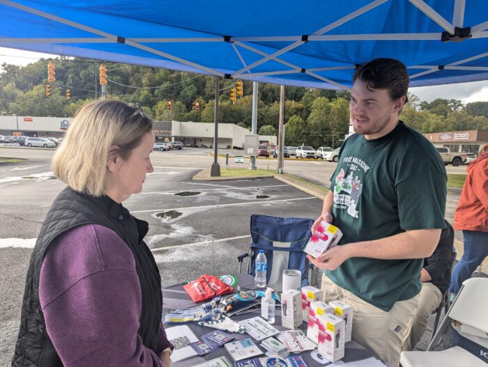 A woman wearing a black vest over a purple shirt stands facing a man wearing a green shirt. He holds a box over a table arrayed with other items. They stand under a blue popup tent in a parking lot.