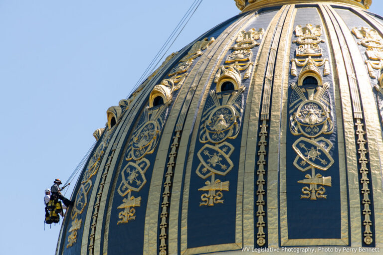 A close up view of the top of the capitol dome. A man scales the side doing repairs. It's a clear blue sky.