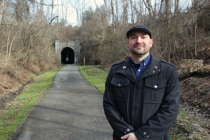 A man in a black cap and black overcoat stands on a gravel path that stretches away behind him towards a tunnel in a hillside.