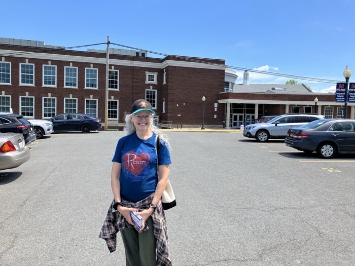 A woman wearing a black baseball cap with loose white hair flowing down over her shoulders and a blue shirt with a red heart and the word "Resist" written in white stands in a parking lot holding a stack of fliers in front of a two story red brick building.