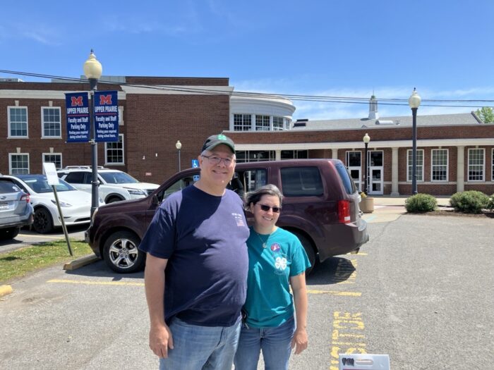 A man wearing a 4H hat and a dark blue t-shirt over jeans stands shoulder to shoulder with a woman wearing a teal 4H shirt and sunglasses with jeans. They stand in the parking lot in front of a two story brick building.