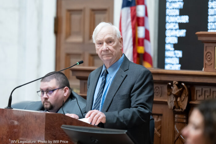 A man in a grey suit over a light blue shirt and dark blue tie stands at a lectern with a microphone pointed at him. Behind him can be seen a larger dark wood dais and flags.