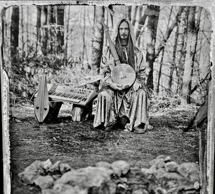 A black-and-white tin type image of a man holding a banjo sitting in the woods.
