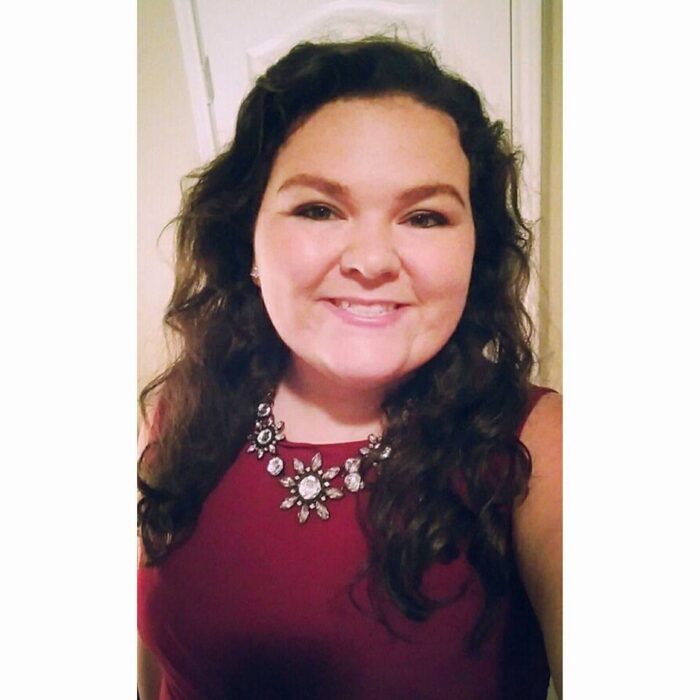 A portrait of a white woman smiling. She is wearing a red shirt and a silver flower necklace. She is smiling.
