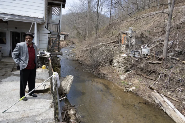 A landscape image of a Black man wearing a suit. He is standing on a concrete wall that sits against a creek and a hill. 