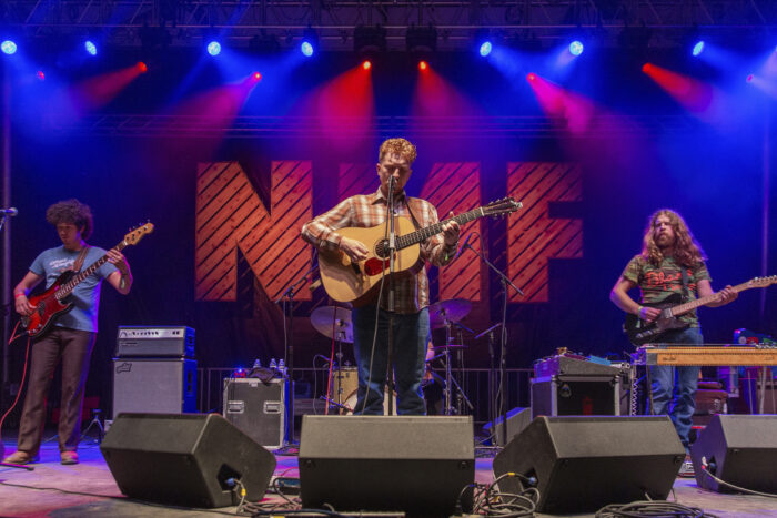 A landscape image of a musician singing center stage. He is holding a guitar. There is one musician on either side of him, and the letters "NMF" are behind him. 