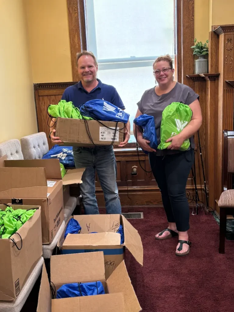 Two adults smile for the camera as they load up WVPB green and blue bags into boxes for Be My Neighbor Day 2025.