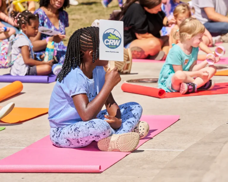 A young girl holds up a fan while she sits on a yoga mat. In the distance are several other children also on yoga mats.