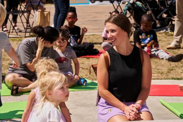 A woman with brown hair looks off the camera and smiles. She sits on a yoga mat with two small children. In the distance are four other people, an adult and three children, who also sit on yoga mats.
