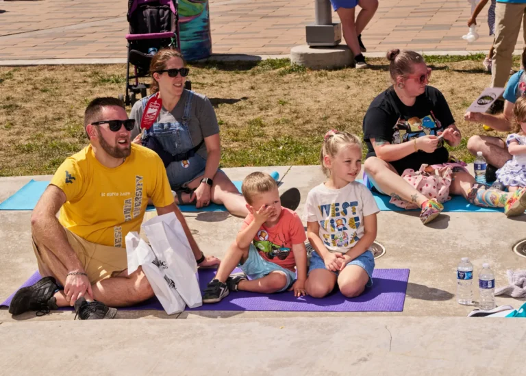 Five people are visible. Two adult women, one adult man, two young children, a boy and a girl. They sit on yoga mats on a sunny day.