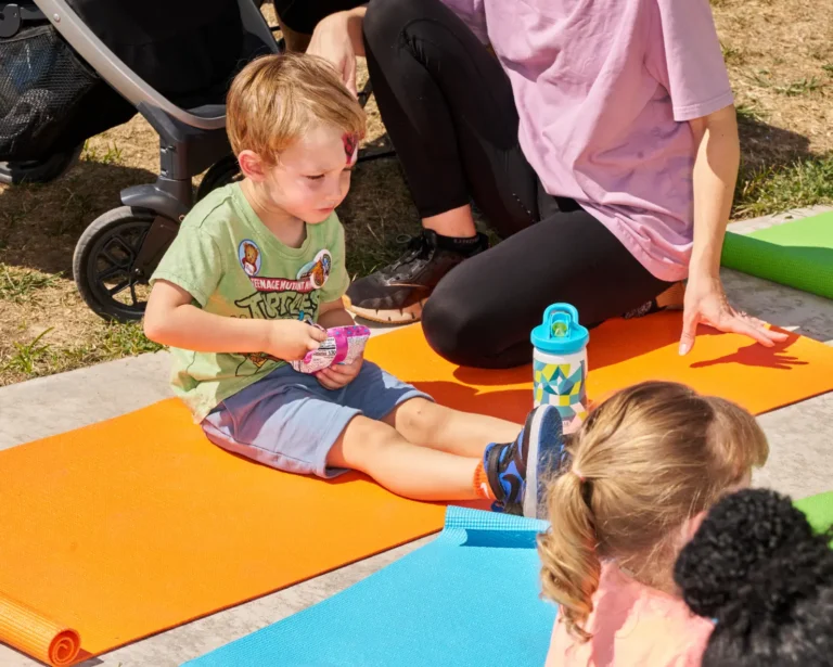 A small boy sits on a yellow yoga mat and eats a snack, outside on a sunny day.