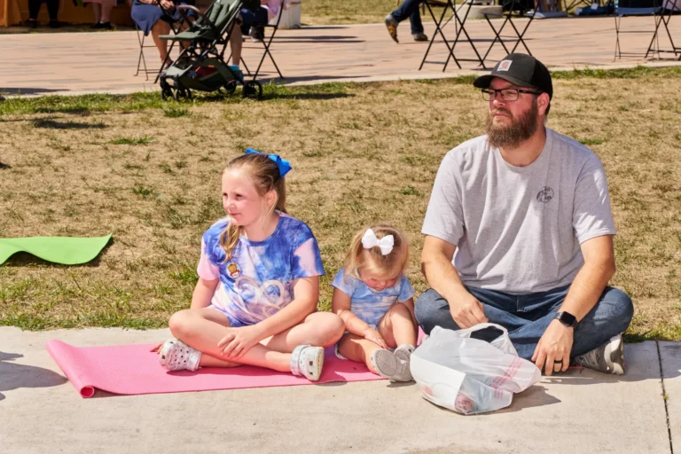 Three people, an adult man and two children, both girls, sit on a yoga mat outside on a sunny day.