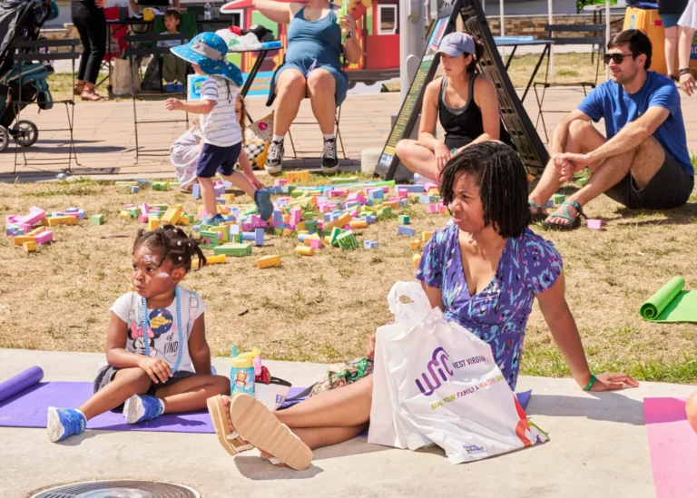 A mother and her daughter sit on a yoga mat. In the background is another family playing with blocks. It is a bright, sunny day.