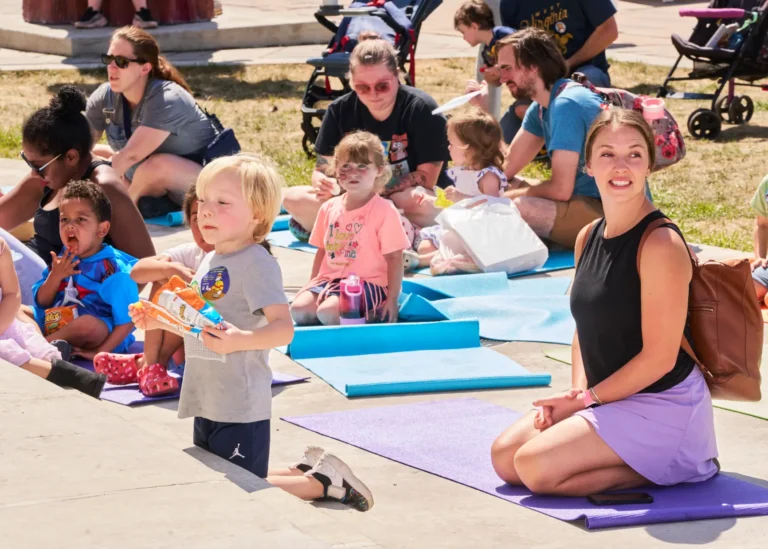 Ten people are visible, five adults and five children. They sit on yoga mats outside on a sunny day.