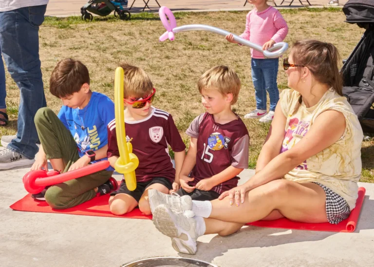 Four people, an adult woman and three young boys, sit on a yoga mat together outside on a sunny day. Two of the boys are holding balloon swords.