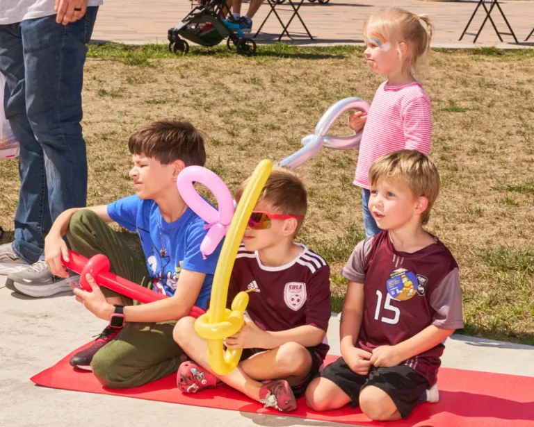 Four children, three boys and a girl, are visible. The boys sit on a yoga mat, and three of the children have balloon swords.
