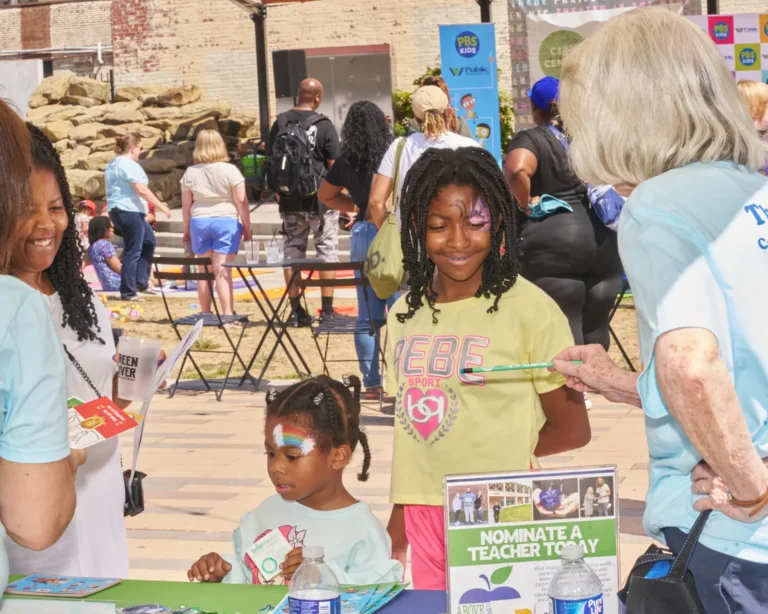 Two young girls stand at a vendor table wearing face paint and smile as they listen to the adults near them talking. One adult in the photo offers one of the girls a green pencil.