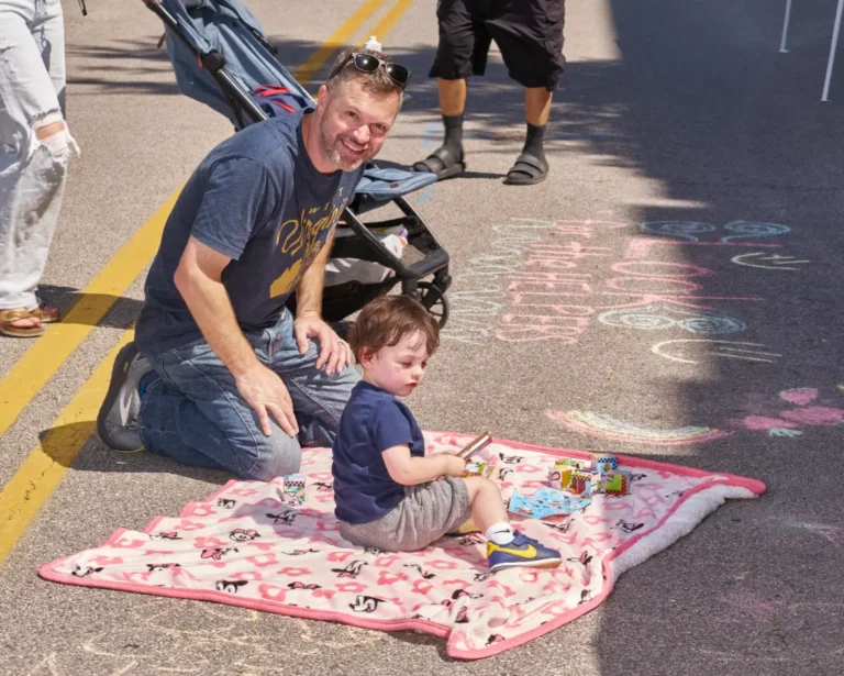 An adult man sits on the ground with a small child who is sitting on a blanket. The man smiles toward the camera as the little boy looks off camera. There is also sidewalk chalk drawings visible.