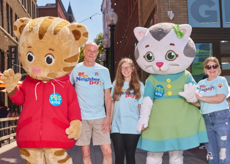 Three adults, a man and two women, stand with Daniel Tiger and Katerina Kittycat on a sunny day.