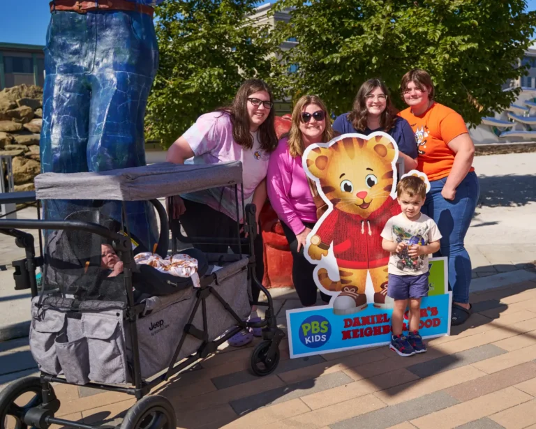 Four adults, all women, and one child, a little boy, pose for a photo around a Daniel Tiger's Neighborhood cutout.