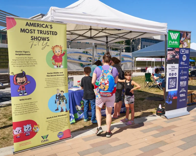 Five people, three adults and two children, are visible standing at a vendor table. A popup banner says, "America's Most Trusted Shows, For You." On the banner are characters such as Daniel Tiger and the Kratt Brothers.