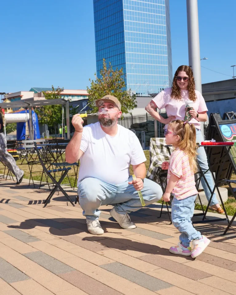 A father blows bubbles, as a small girl watches. A woman in the background is watching them, smiling.