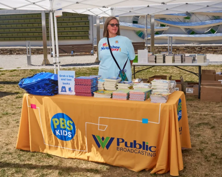 An adult woman stands behind a vendor table. The table has a tablecloth with the WVPB and PBS logos. On the table are stacks of books.