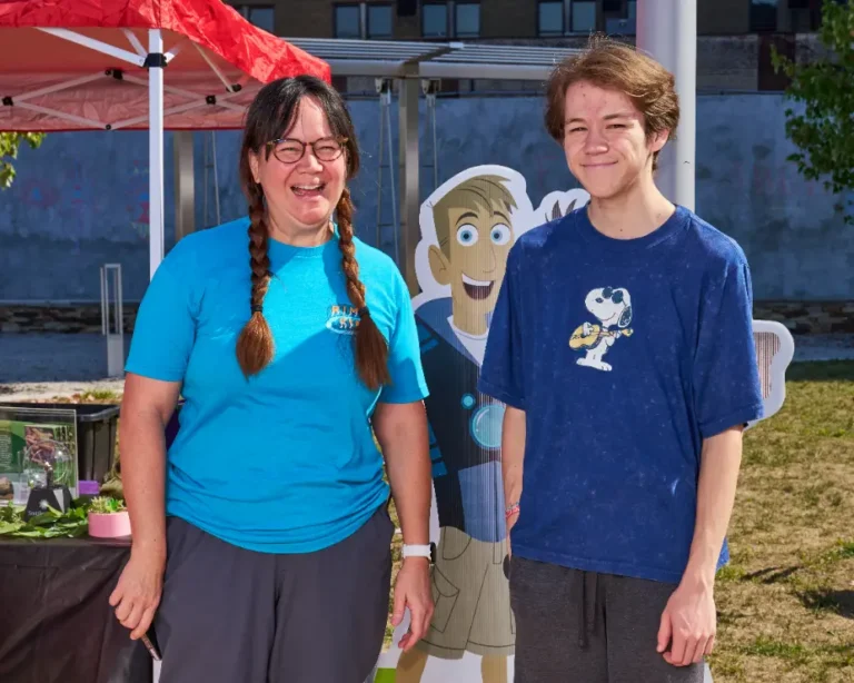 A woman wearing glasses, whose hair is braided into pigtails smiles for the camera. Next to her is a young man wearing a blue shirt with Snoopy on it.