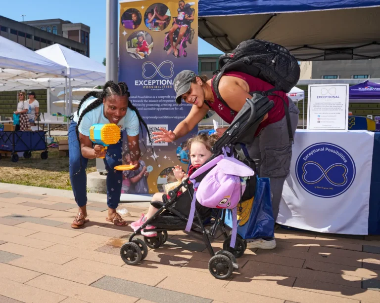 Two adults, both women, and one child are visible. The child is sitting in a stroller. One woman is holding a bubble blower and is cheering. The other adult is looking at the child cheering.