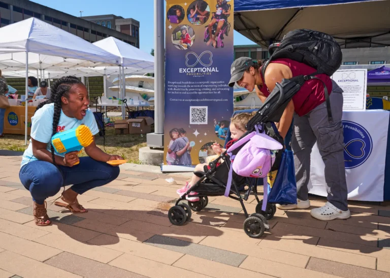 Two adults, both women, and one child are visible. The child is sitting in a stroller. One woman is holding a bubble blower and is cheering. The other adult is looking at the child cheering.