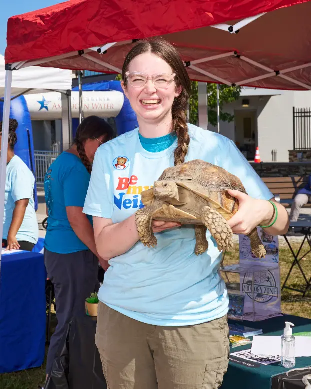 A woman wearing glasses and with a single braid smiles for the camera. She is holding a large tortoise.