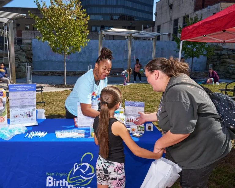 Two adult women and one young girl stand at a vendor table. The vendor is handing swag to the girl.