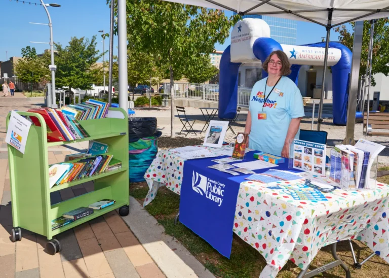 An adult woman stands at a table outside on a sunny day. There are books on her table in a book rack nearby. A tablecloth reads, "Kanawha County Public Library."