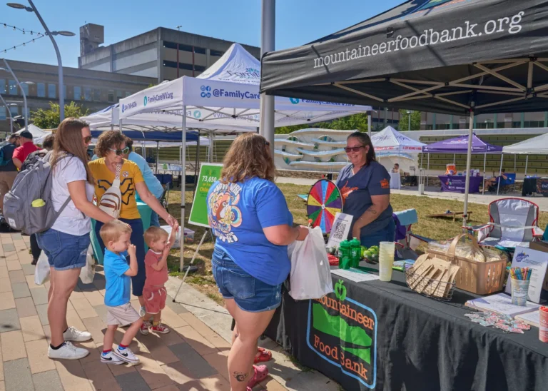 Four adults are visible and two small children. There is a vendor table with a tablecloth that reads, "Mountaineer Food Bank."