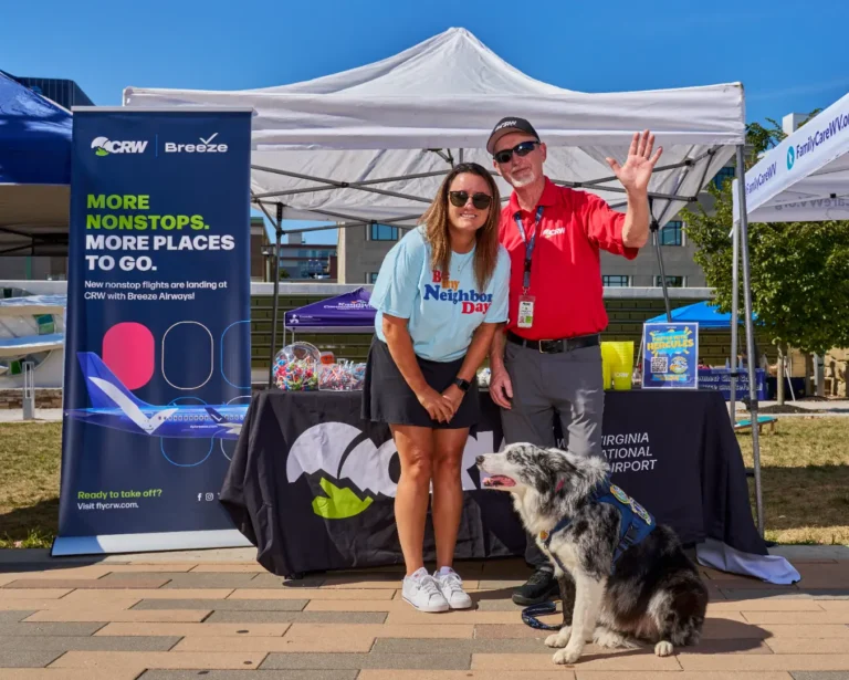 Two adults stand in front of a vendor table. There is a popup banner that reads, "More Nonstops. More places to go. CRW, Breeze." With the adults is a dog wearing a harness.
