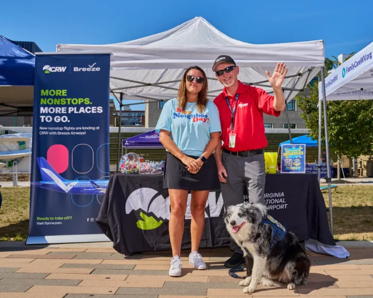 Two adults stand in front of a vendor table. There is a popup banner that reads, "More Nonstops. More places to go. CRW, Breeze." With the adults is a dog wearing a harness.