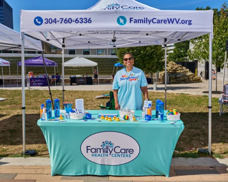 A woman stands behind a vendor table. There is a blue tablecloth on the table with a logo that reads, "Family Care Health Centers." A popup tent is above her with a website that reads, familycarewv.org, 304-760-6336.