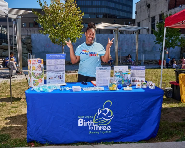 A woman stands at a vendor table, holding up her hands in two peace signs. The tablecloth on her table reads, "West Virginia Birth to Three, Growing Together."