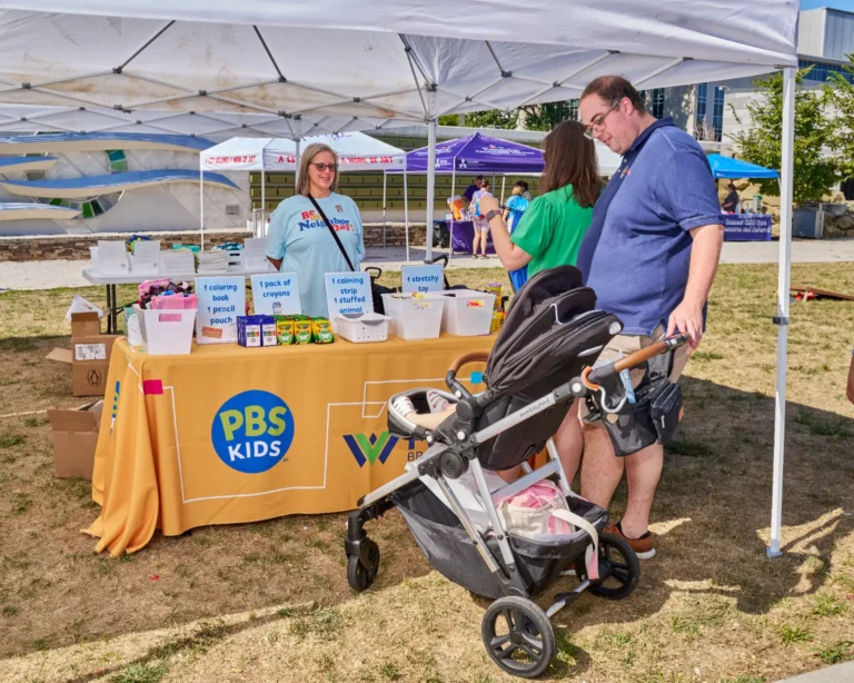 Three adults, two women and one man. A stroller is also visible with a child sitting inside. There is a table visible with the PBS and WVPB logos.