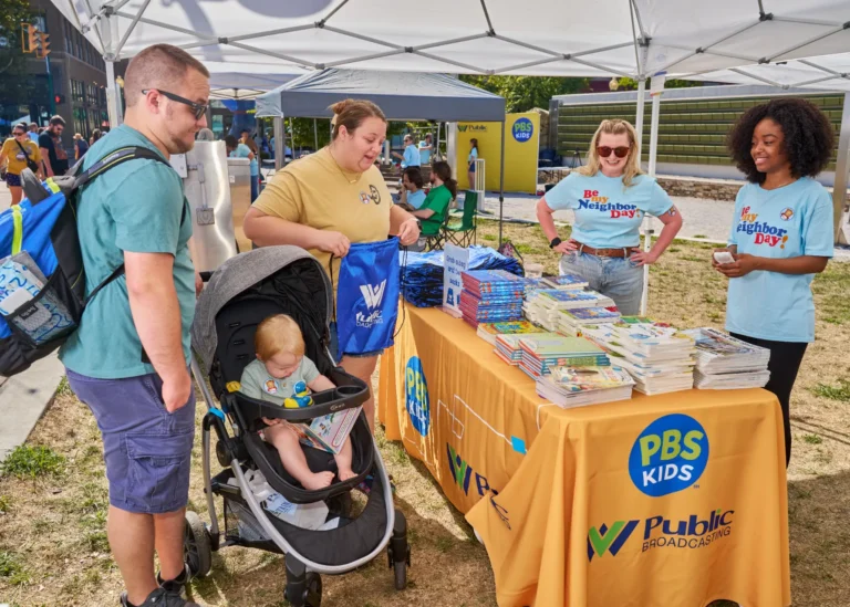 Four adults, one child in a stroller. A tablecloth is visible and stacked with books. The PBS and WVPB logos are visible on a tablecloth.