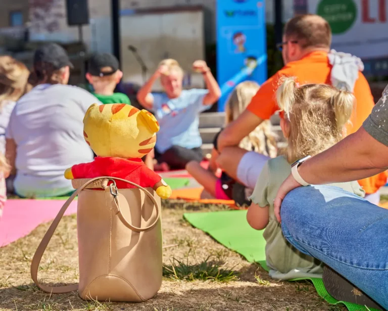 A Daniel Tiger stuffed toy has been placed inside of a mother's purse. The mother's arm is only visible, and she sits with a small girl with blond hair and pigtails. They sit on a green yoga mat.