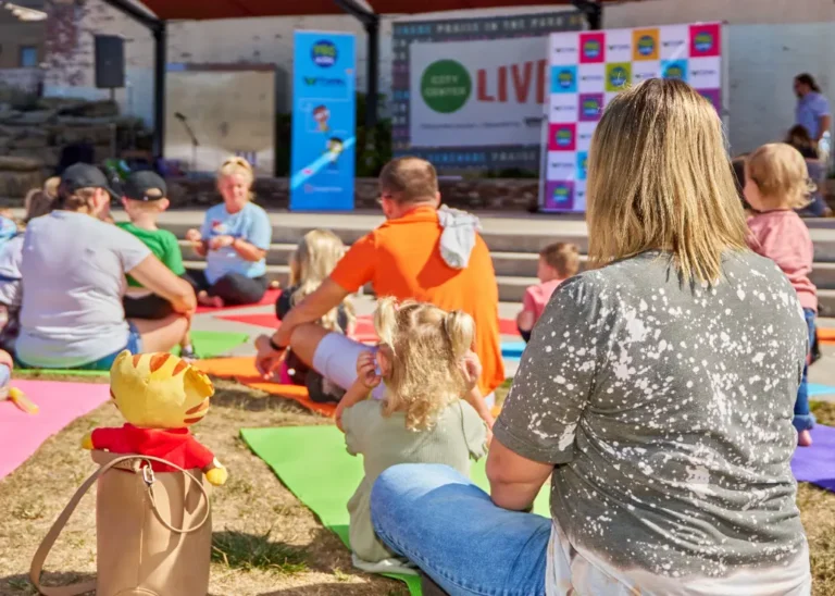 Several people with their backs to the camera. They are sitting on yoga mats outside on a sunny day. Near the camera is a purse with a small Daniel Tiger stuffed toy inside.